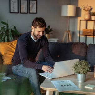 Eine Person sitzt auf dem Sofa am COuchtisch und tippt etwas in das geöffnete Laptop ein.