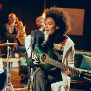 Eine Person sitzt in einem kleinen Club mit ihrer Gitarre auf einem Hocker und singt in ein Mikrofon.
