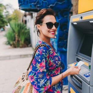 Eine Frau steht lächelnd vor einem Geldautomaten und hält eine Kreditkarte in der Hand