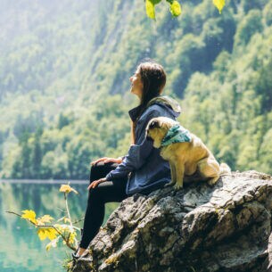 Eine Person und ein kleiner Hund sitzten zusammen auf einem Felsen vor einem See, der von Wald umsäumt ist
