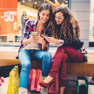 Zwei Personen sitzen in einer Shoppingmall auf einer Bank und schauen gemeinsam auf ein Smartphone und eine Kreditkarte.
