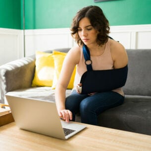 Eine Frau mit ruhiggestelltem Arm sitzt auf einem Sofa und tippt mit der freien Hand auf ihrem Laptop.