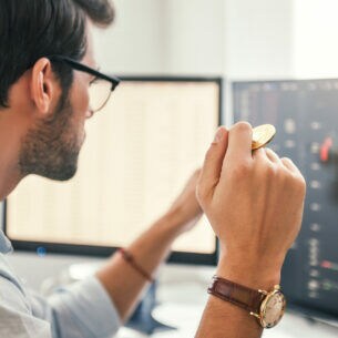 Ein Mann schaut auf den Kursverlauf auf seinem Computermonitor und hält eine Münze in der Hand