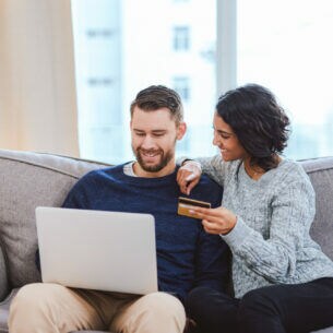 Eine Frau und ein Mann sitzen auf einem Sofa. Die Frau hält eine Kreditkarte in der Hand, der Mann hat einen Laptop auf dem Schoß.