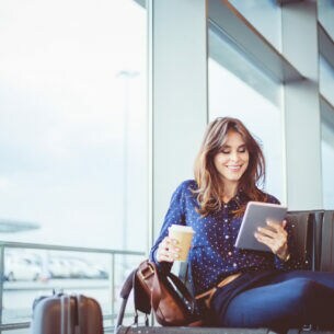 Eine Person sitzt am Flughafen und hält einen Kaffeebecher und ein Tablet in den Händen. Ein Koffer steht neben ihr