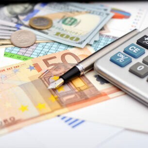 Ein Taschenrechner, ein Füllfederhalter, Banknoten und Münzen in verschiedenen Währungen liegen auf Zetteln mit Tabellen und Zahlen.