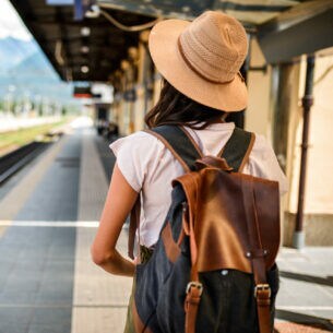 Eine Person in Rückansicht mit Rucksack und Hut steht am Bahnsteig.