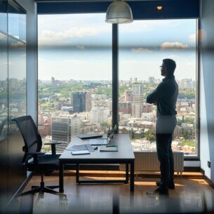 Eine Person steht in einem Büro vor einer Fensterfront und blickt über das Stadtpanorama.