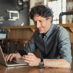Eine Person blickt auf sein Smartphone und sitzt vor einem geöffneten Laptop.