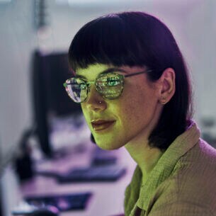 Portraitaufnahme einer Frau mit Brille, in deren Gläsern sich die Schrift eines Computerbildschirms widerspiegelt.