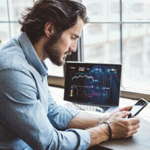 Ein Mann im hellblauen Hemd an einem Schreibtisch blickt auf das Smartphone in seiner Hand. Auf dem Tisch steht ein Notebook mit Börsenkursen auf dem Bildschirm.