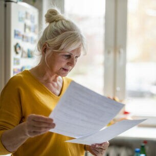 Eine Frau im gelben Pullover steht in einer Küche am Fenster. Sie blickt auf Dokumente, die sie in den Händen hält.