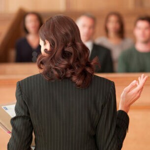 Eine Person im Gerichtssaal mit langen, rotbraunen Haaren in Nadelstreifenoberteil steht mit Dokumenten in einer Hand Personen gegenüber, die in den Gerichtssaalreihen sitzen.