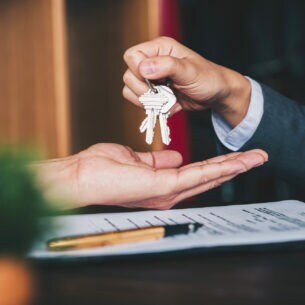 Eine Person hält einen Schlüsselbund über die aufgehaltene Hand einer anderen Person, darunter liegen ein Vertrag und ein Füller.