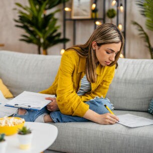 Eine Frau mit gelber Hemdjacke sitzt auf einem Sofa und füllt Dokumente aus.