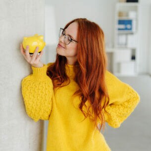 Eine Person mit gelbem Pullover hält ein gelbes Sparschwein in der Hand und schaut es direkt an.