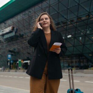 Eine Frau vor einem verglasten, modernen Gebäude hält mit einer Hand ein Telefon ans Ohr, in der anderen hält sie Reisedokumente. Neben ihr steht ein Koffer.