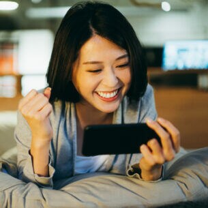 Eine Person liegt auf einem Bett und schaut lächelnd auf das Smartphone, das sie in einer Hand hält.