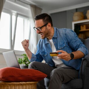 Ein Mann sitzt auf einem Sofa vor einem aufgeklappten Laptop, der auf einem Hocker steht. In einer Hand hält er eine Geldkarte, die andere ballt er zu einer Faust.