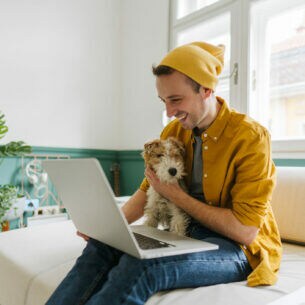 Eine Person sitzt auf einem Sofa, krault einen Hund auf dem Schoß und blickt in ein geöffnetes Notebook.