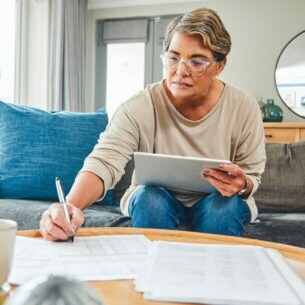 Eine Person sitzt mit einem Tablet auf den Knien auf einem Sofa. Sie beugt sich mit einem Stift in der Hand nach vorne zu Unterlagen auf dem Couchtisch.