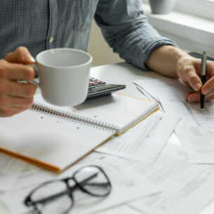 Eine Person am Tisch hält eine Tasse und einen Kugelschreiber in Händen. Auf dem Tisch liegen Dokumente, ein Taschenrechner und eine Brille.