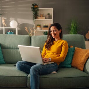 Eine Person sitzt auf einem Sofa und schaut auf das geöffnete Notebook vor sich.