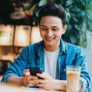 Eine Person sitzt mit einem Café vor sich am Tisch und blickt auf das Smartphone, das sie in den Händen hält.