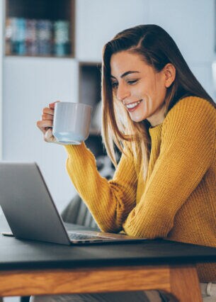 Eine junge Frau sitzt lächelnd mit einer Tasse vor ihrem Laptop