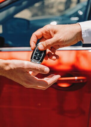 Eine Hand übergibt einen Autoschlüssel in eine andere Hand. Im Hintergrund ist ein rotes Auto zu sehen