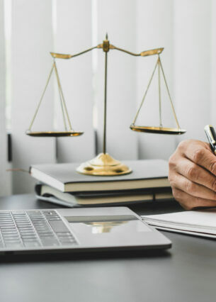 Person im Anzug mit Laptop, Block und Stift bei der Arbeit am Schreibtisch, im Hintergrund steht die Waage der Gerechtigkeit