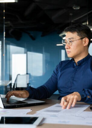 Eine Person sitzt im Büro an einem Schreibtisch mit zahlreichen Dokumenten und blick auf ein geöffnetes Laptop