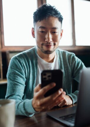 Eine Person sitzt an einem Tisch, auf dem ein geöffneter Laptop steht, hält ein Smartphone in der Hand und blickt aufs Display