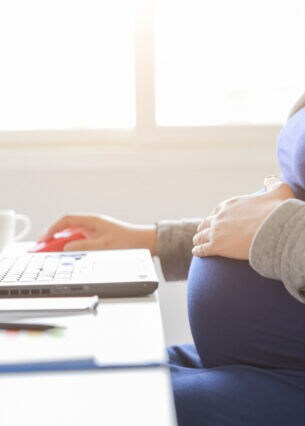 Eine schwangere Frau sitzt an einem Tisch vor einem aufgeklappten Notebook. Eine Hand hat sie an der Computermaus, die andere auf dem Babybauch.