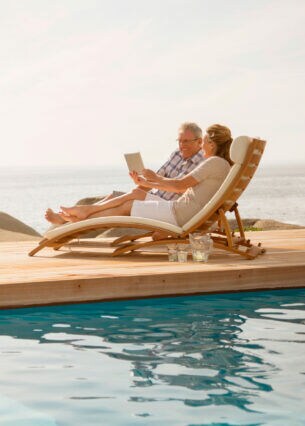 Zwei ältere Personen liegen auf einer Holzliege am Pool, im Hintergrund ist das Meer zu sehen