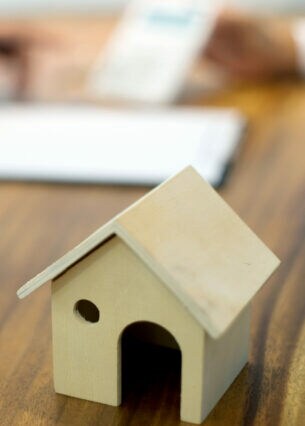 Ein kleines Hausmodell aus Holz steht auf einem Schreibtisch, im Hintergrund sind unscharf zwei Personen und Unterlagen zu erkennen.
