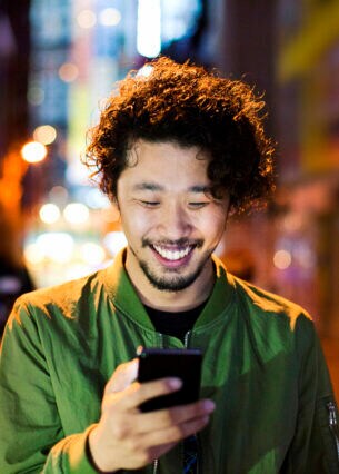 Ein Mann mit Smartphone in der Hand, in dessen Richtung er lächelt. Im Hintergrund eine verschwommene Großstadtkulisse.