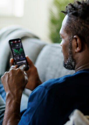 Eine Person sitzt auf einem Sofa und blickt auf sein Smartphone, auf dem Aktienkurse zu sehen sind.
