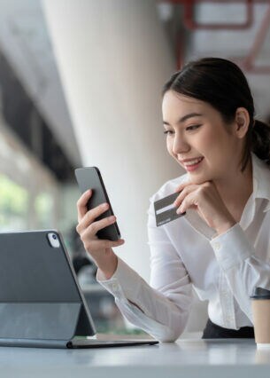 Eine lächelnde Person schaut auf das Smartphone, das sie in der Hand hält. In der anderen Hand hält sie eine Kreditkarte. Vor ihr stehen ein Tablet und ein Becher.