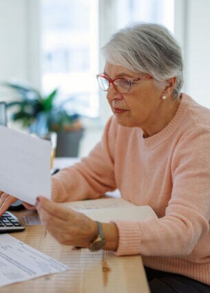 Eine ältere Dame an einem Tisch hält ein Blatt Papier in der Hand und tippt auf einen Taschenrechner.