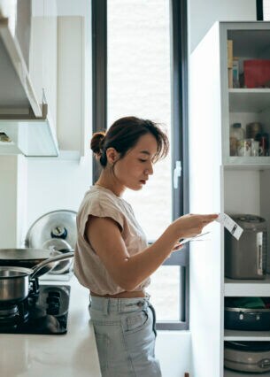 Eine junge Frau steht in einer Küche. Sie sieht sich einen Zettel an, den sie in der Hand hält.