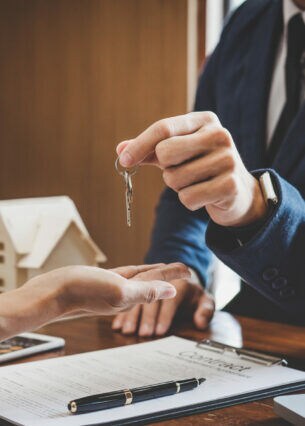 Eine Person in Anzug übergibt einen Schlüssel in eine ausgestreckte Hand. Auf dem Schreibtisch steht im Hintergrund ein kleines Holzhaus