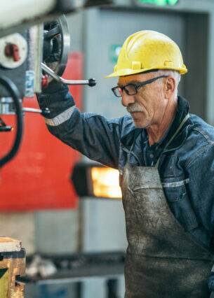Ein älterer Mann mit gelbem Helm und Blaumann steht an einer Industriebohrmaschine.