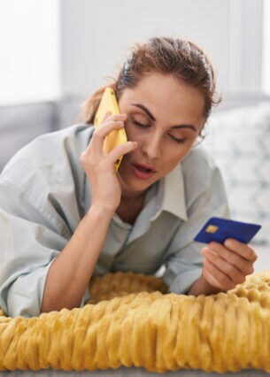Eine Person liegt bäuchlings und telefonierend auf dem Sofa und hält eine Kreditkarte in der Hand.