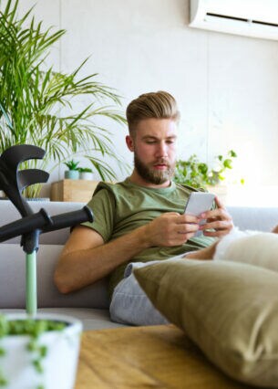 Ein Mann liegt auf dem Sofa und hält ein Smartphone in der Hand. Er hat seinen eingegipsten Fuß hochgelegt.