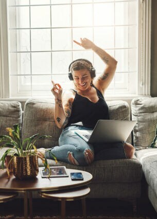 Eine Person sitzt mit Kopfhörern und einem Laptop auf dem Schoss auf einem Sofa und wirft lächelnd die Arme in die Höhe
