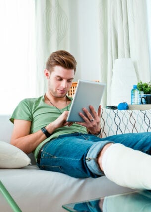 Eine Person mit gebrochenem Bein sitzt auf einem Sofa und schaut auf sein Tablet, neben ihr stehen Krücken.