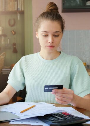 Eine Person sitzt am Küchentisch, schaut auf ihre Bankkarte und hat mehrere Dokumente vor sich liegen sowie die andere Hand auf einem geöffneten Laptop.