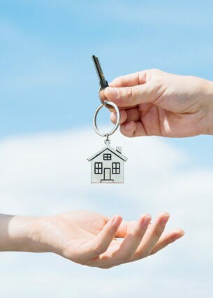 Übergabe eines Schlüssels mit metallenem Schlüsselanhänger in Form eines Hauses, Nahaufnahme der Hände vor blauem Himmel.