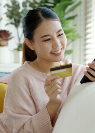 Eine Person sitzt auf einem Sofa, halt eine Bankkarte in der einen Hand und blickt auf ihr Smartphone in der anderen Hand.